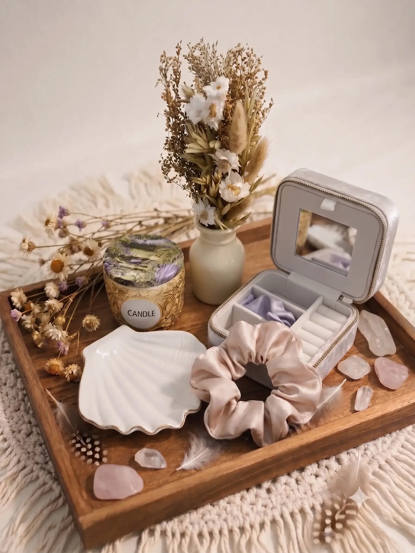 A meticulously curated boho-chic vanity decor set arranged on a smooth solid wood tray, featuring a silver-grey velvet jewelry box with an open mirror, a white ceramic shell ring dish with a gold rim, a blush satin scrunchie, a scented candle in an embossed gold jar, a minimalist cream vase with a preserved dried floral bouquet, scattered rose quartz decorative crystals, and feathers.